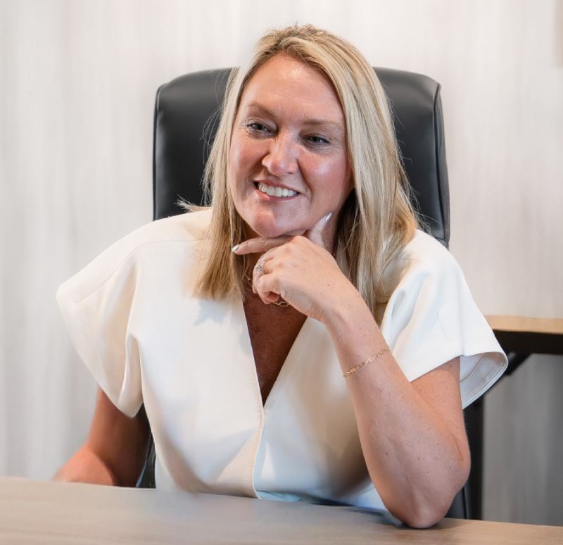 A smiling Herringbone leader in a white top seated at a desk, chin resting on her hand