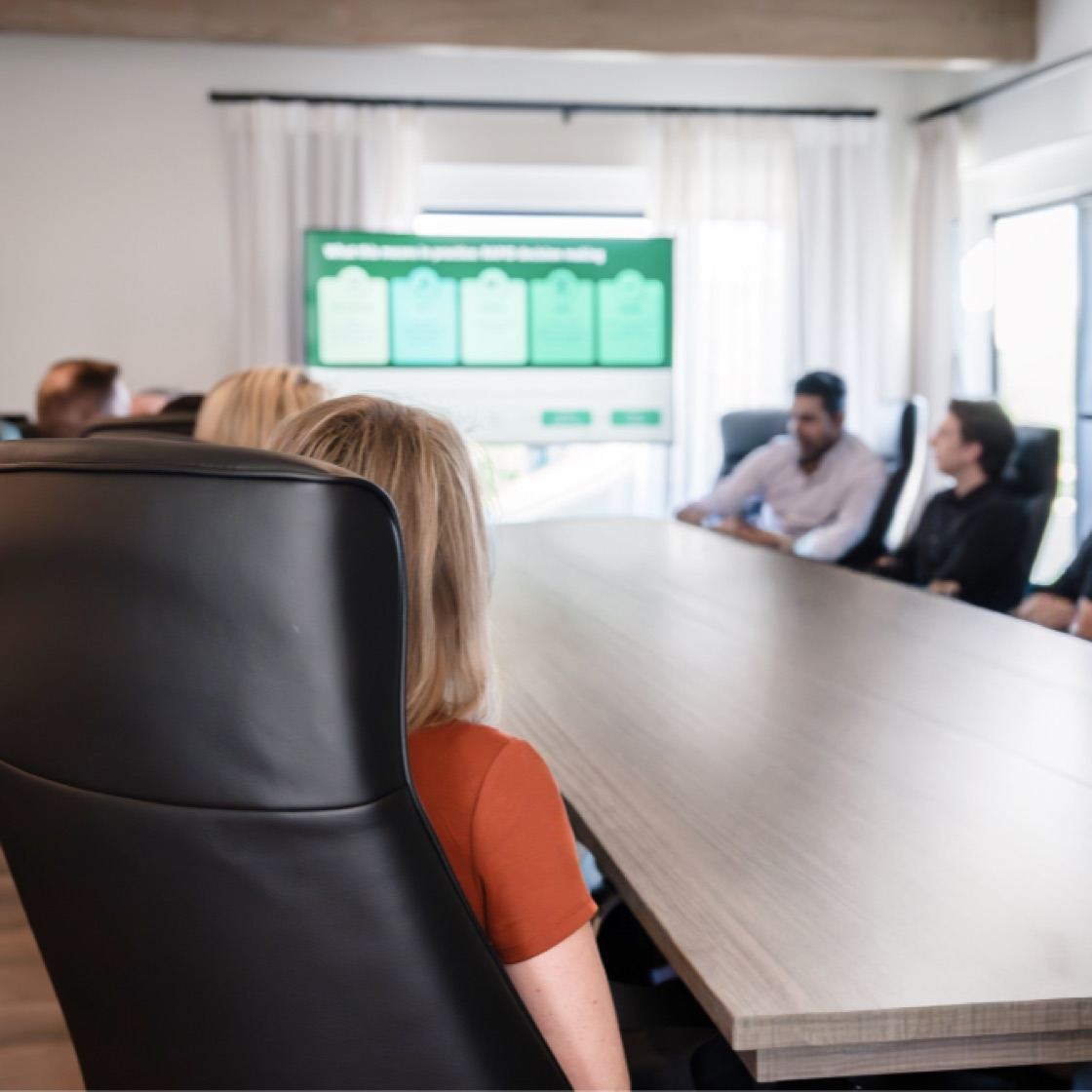 Herringbone team seated around a long wooden conference table during a working session, a presentation on the screen at the far end of the room