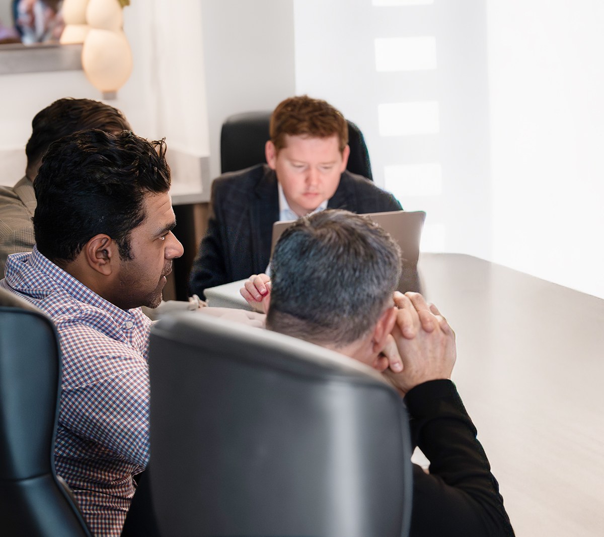Herringbone team in a working session at a conference table: a man in a checked shirt talks with a colleague resting his chin on clasped hands while another works on a laptop at the head of the table