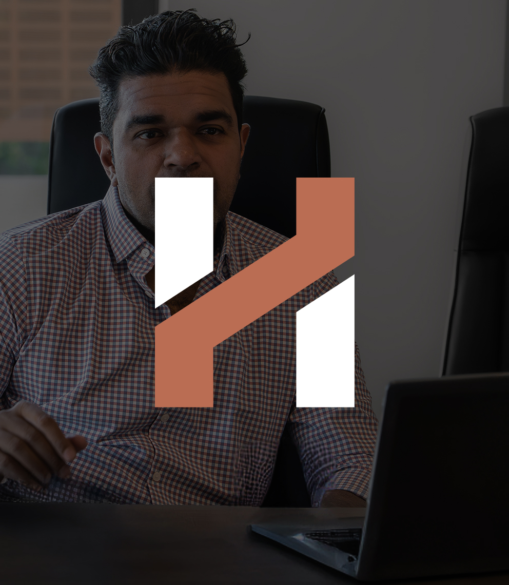 A Herringbone team member in a checked shirt at a desk with a laptop, under a dark overlay with the Herringbone H mark